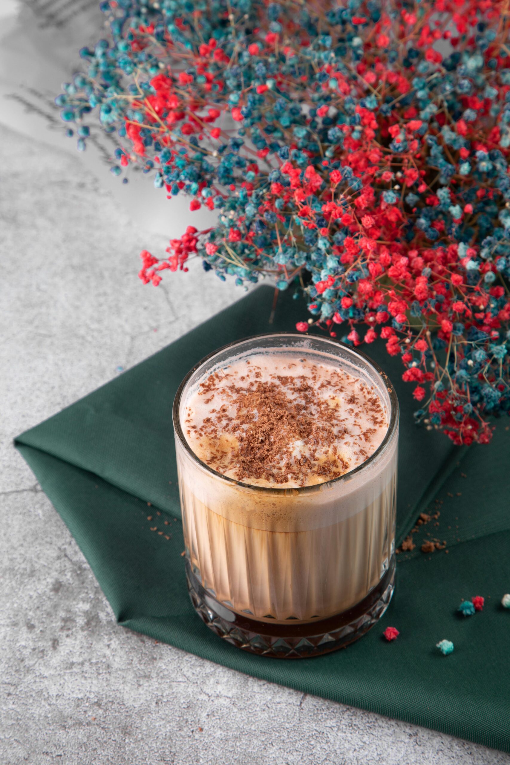 Glass of hot chocolate on green napkin with colorful flowers for a cozy vibe.